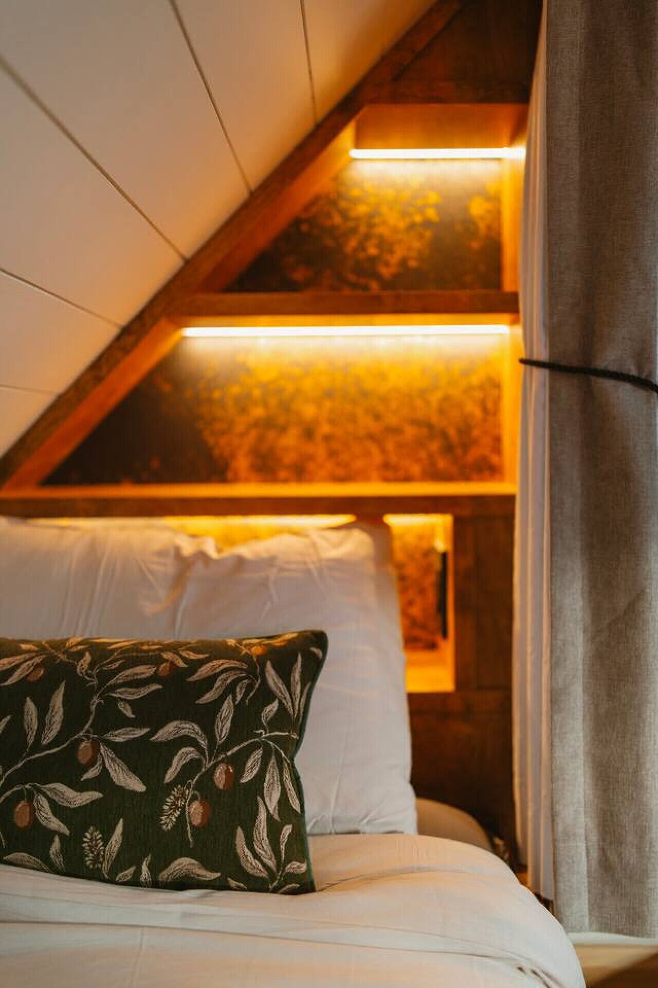 Une chambre confortable avec un plafond en pente, un lit recouvert de draps blancs, un coussin à motifs verts et une vue sur une forêt par la fenêtre.