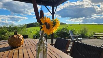 A wooden table with a glass bottle and a sunflower, a carved pumpkin, and outdoor furniture under a covered area with a view of a green field.