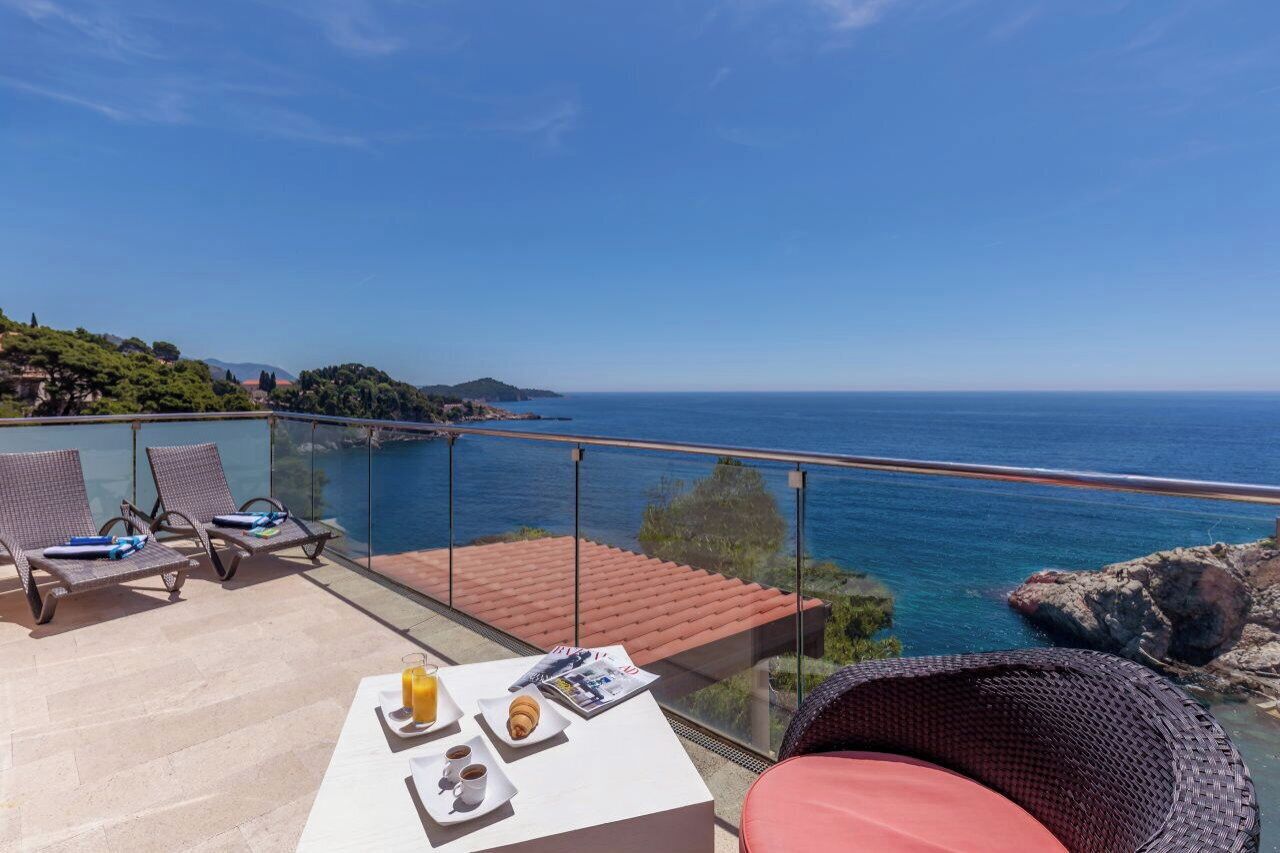 A balcony with a glass railing overlooking a body of water, lounge chairs, and a table with breakfast items.