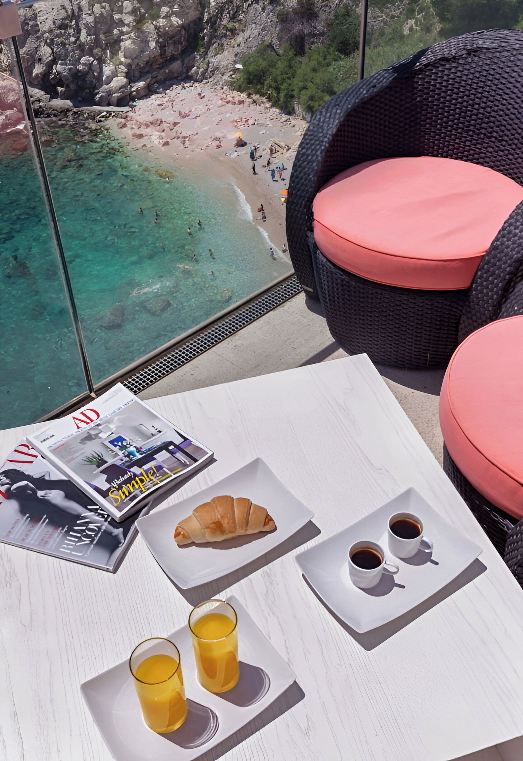 Un tavolo con prodotti per la colazione, una rivista e vista su una spiaggia e scogliere rocciose.