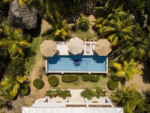 Vista aérea de un resort con una piscina rectangular, rodeada de palmeras y una estructura con techo de paja.