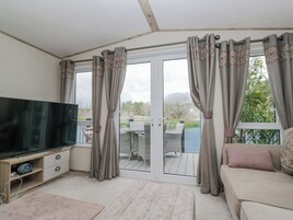 Quarto com televisão, sofá, porta de vidro deslizante para área externa com mesa e cadeiras, e vista para árvores e um corpo d'água.