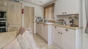A kitchen with wooden cabinets, a sink, a stove, and a dining area with a chair. - Carp Lodge (Carnforth)