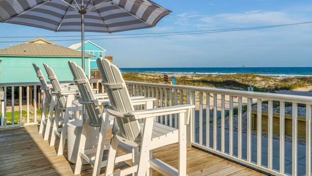 Une terrasse en bois, agrémentée de chaises Adirondack blanches et d’un parasol rayé, offre une vue sur la plage.