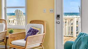 Une pièce avec un fauteuil en osier, une horloge au mur et une vue sur la plage par la porte.