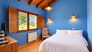 Un dormitorio con una cama, una silla, un escritorio con un modelo de barco, un radiador y dos ventanas con contraventanas de madera.