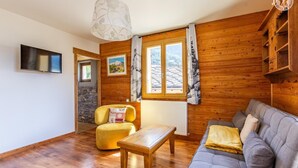 Ein gemütliches Wohnzimmer mit Holzwänden, einer Couch, einem gelben Sessel, einem Holztisch, einem Fernseher und einem Fenster mit Blick auf einen Berg.