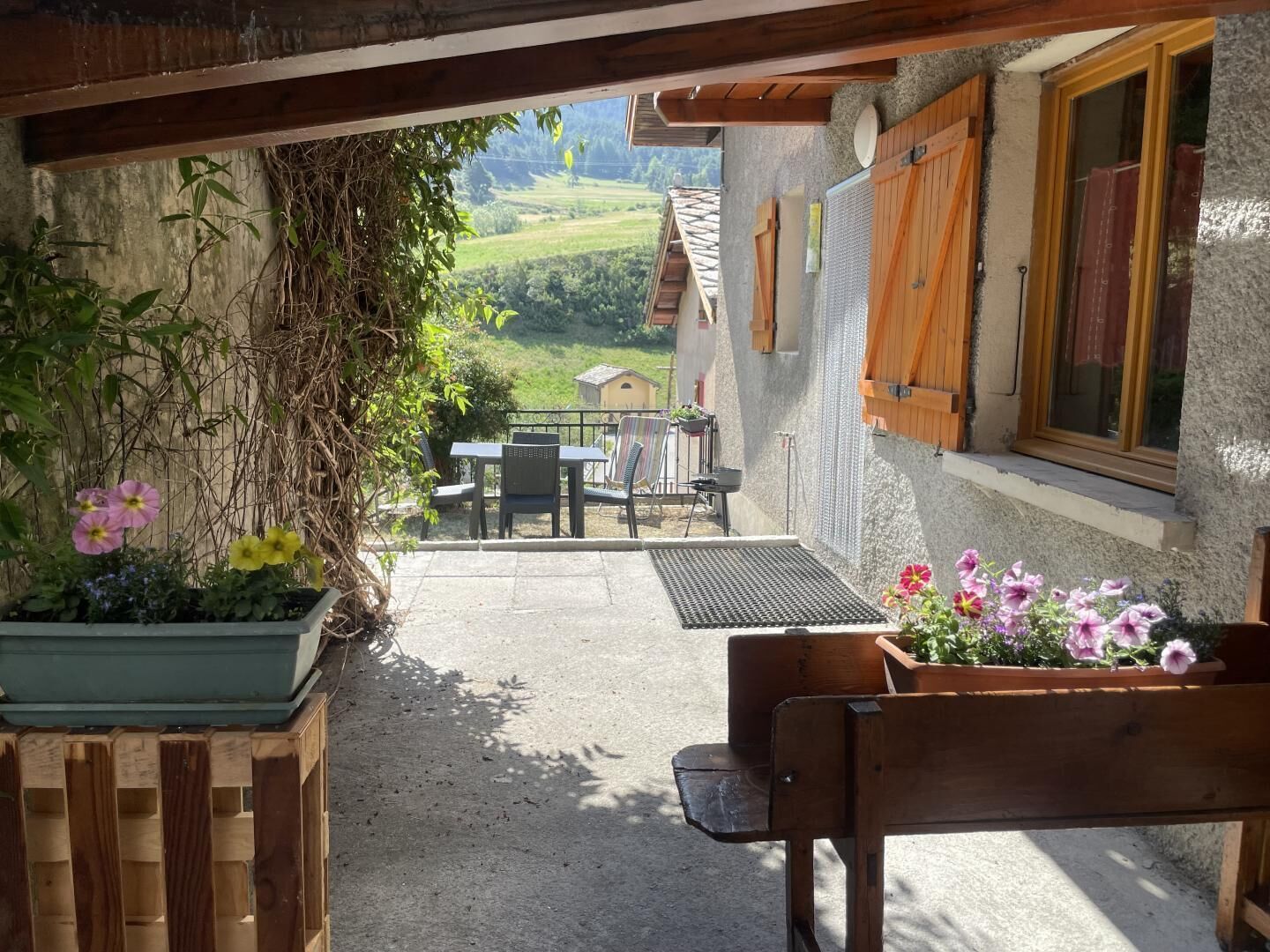 A patio with a wooden bench, a table, and chairs, surrounded by potted plants and a view of a rural landscape.