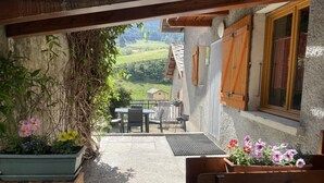 Eine Terrasse mit einer Holzbank, einem Tisch und Stühlen, umgeben von Blumentöpfen und Blick auf eine ländliche Landschaft.
