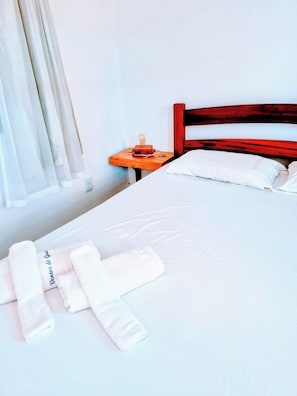 A neatly made bed with white linens, a wooden nightstand with a lamp, and a window with sheer curtains. - Pousada Ventos do Guajiru  (Itarema)