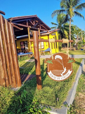 A rustic building with a yellow exterior, a wooden structure with a roof, and a sign with palm trees and text.