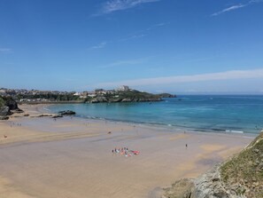 Uma praia costeira com céu claro, faixa de areia e edifícios ao fundo.