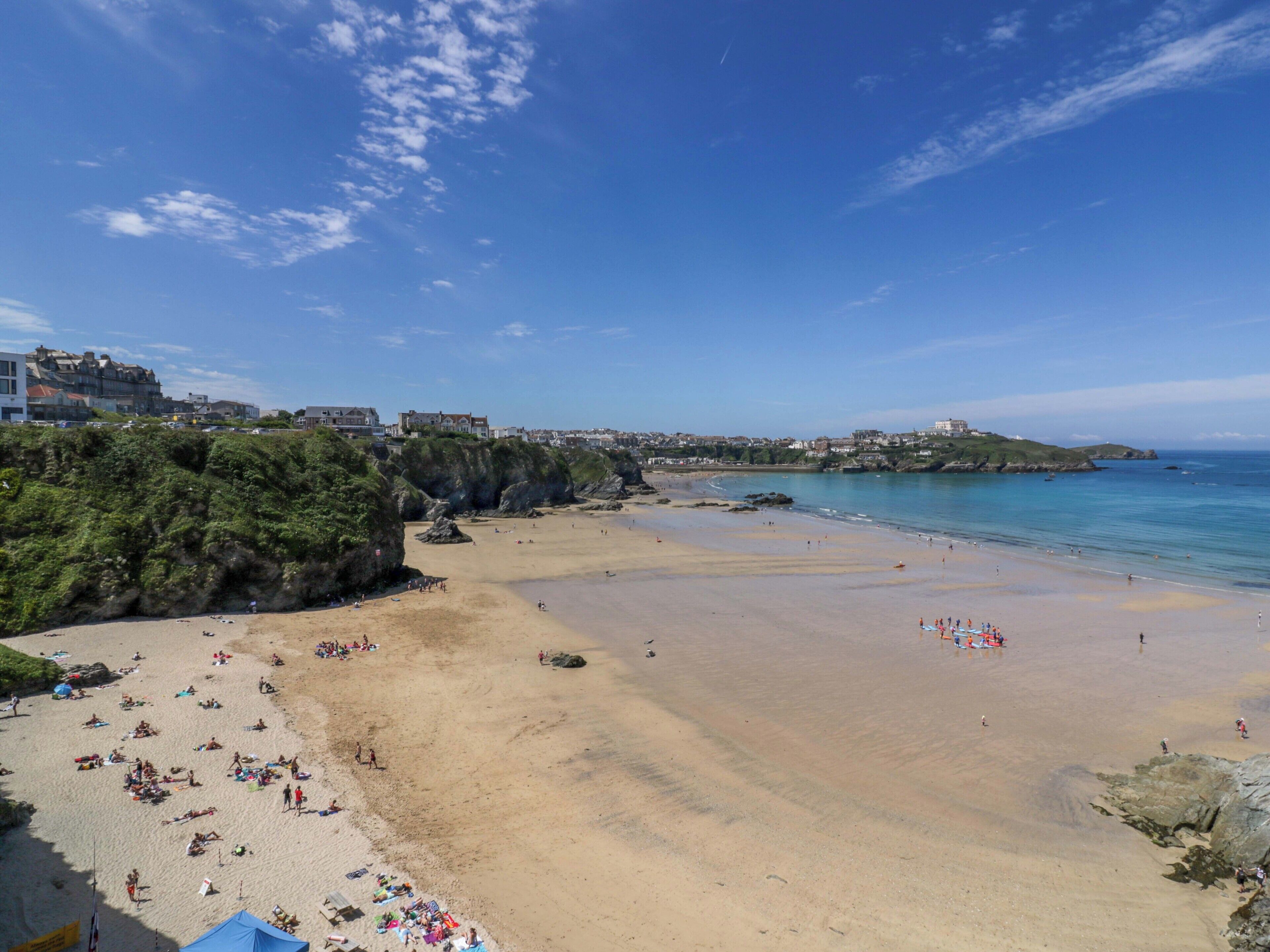 A beach with people sunbathing and swimming, a rocky coastline, and buildings in the background.