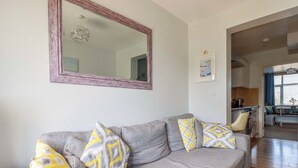 A living room with a grey sofa, patterned cushions, a large mirror, and a view into the kitchen area. - Tolcarne Reach (Newquay)