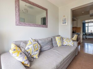 Ein Wohnzimmer mit grauem Sofa, gemusterten Kissen, einem großen Spiegel und Blick in den Küchenbereich.