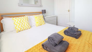 Uma cama bem arrumada com almofadas estampadas, uma mesa de cabeceira com abajur, e uma porta ao fundo.