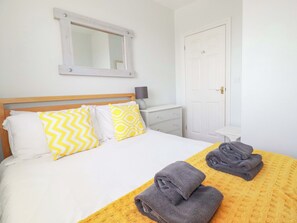 Uma cama bem arrumada com almofadas estampadas, uma mesa de cabeceira com abajur, e uma porta ao fundo.