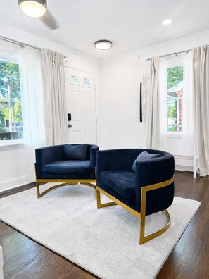 Una habitación con dos sillas azul oscuro de marcos dorados, un tapete blanco, una puerta blanca con paneles de vidrio y una ventana con cortinas.