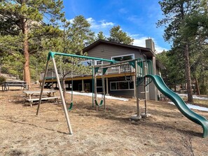 Ein Spielplatz mit Schaukeln und Rutsche, umgeben von Bäumen und einem Gebäude mit Veranda.