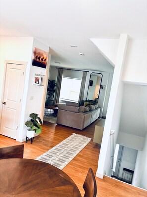 Ein modernes Wohnzimmer mit einer Couch, einem Esstisch mit Stühlen, einem großen Fenster und einer Zimmerpflanze.