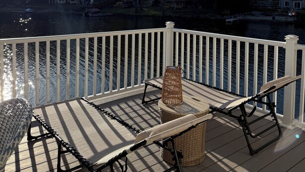 A wooden deck with a white railing, a wicker chair, a table, and a view of houses and trees across a body of water. - Cozy Lake Cottage in charming Dunstable (Dunstable)