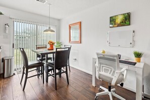 A modern kitchen with a dining area, a white desk with a computer, and a large window with blinds. - The Oaks at Burnell – Private Houston Home (Houston)