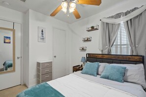 Een slaapkamer met een bed, een plafondventilator, een nachtkastje, een spiegel, een kledingkast en een raam met gordijnen.