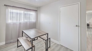 Ein Zimmer mit Holztisch, einer Bank, einer weißen Tür und einem Fenster mit durchsichtigen Vorhängen.