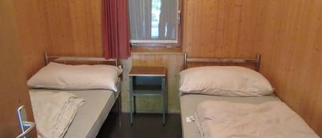 A small, single-room accommodation with two beds, a small table, and a window with a curtain.