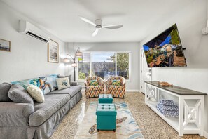 Una sala de estar con un sofá gris, cojines estampados, un reposapiés color turquesa, un ventilador de techo y un ventanal con persianas.