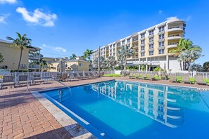A swimming pool with lounge chairs and a white fence, surrounded by palm trees and a multi-story hotel building. - 🌴 Crabby Bungalow | Duplex Unit, Walk to Crescent Beach, & Hot Tub (Siesta Key)