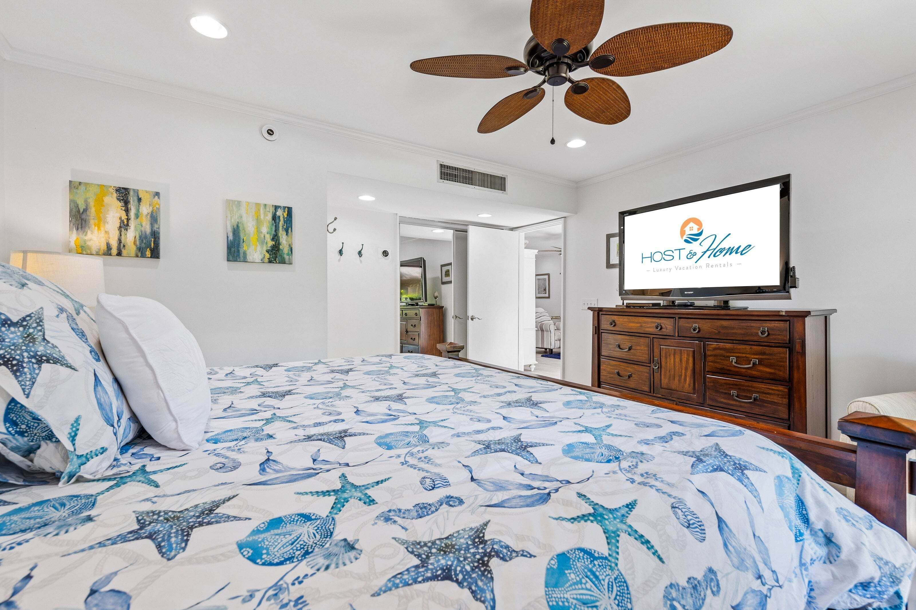 A bedroom with a bed, a ceiling fan, a TV, and artwork on the wall.