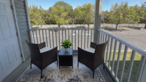 Un balcon fermé doté de deux fauteuils en osier et d’une petite table, offrant une vue sur un parking et des arbres.