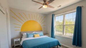 Une chambre à coucher comprenant un lit, une table de chevet, un ventilateur de plafond, une fenêtre avec des rideaux et une fresque représentant un soleil sur le mur.