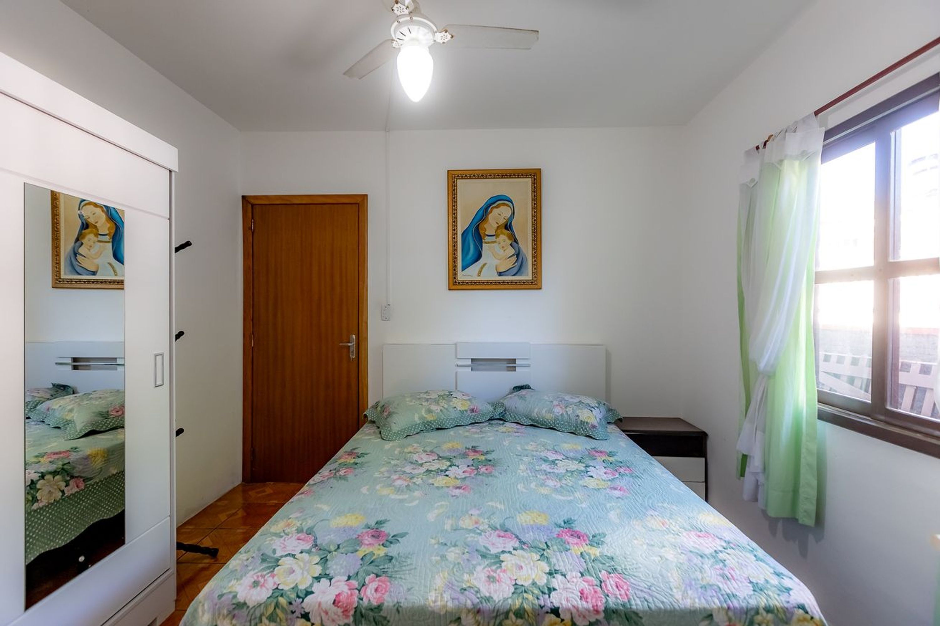Ein Schlafzimmer mit Bett, einem Kleiderschrank, einer Tür, einem Deckenventilator und zwei gerahmten Bildern an der Wand.