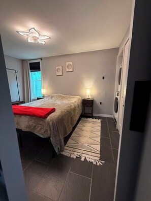 Ein Schlafzimmer mit einem Bett, Nachttischlampen, einem Teppich, einem Fenster mit Vorhängen und einer Waschmaschine.