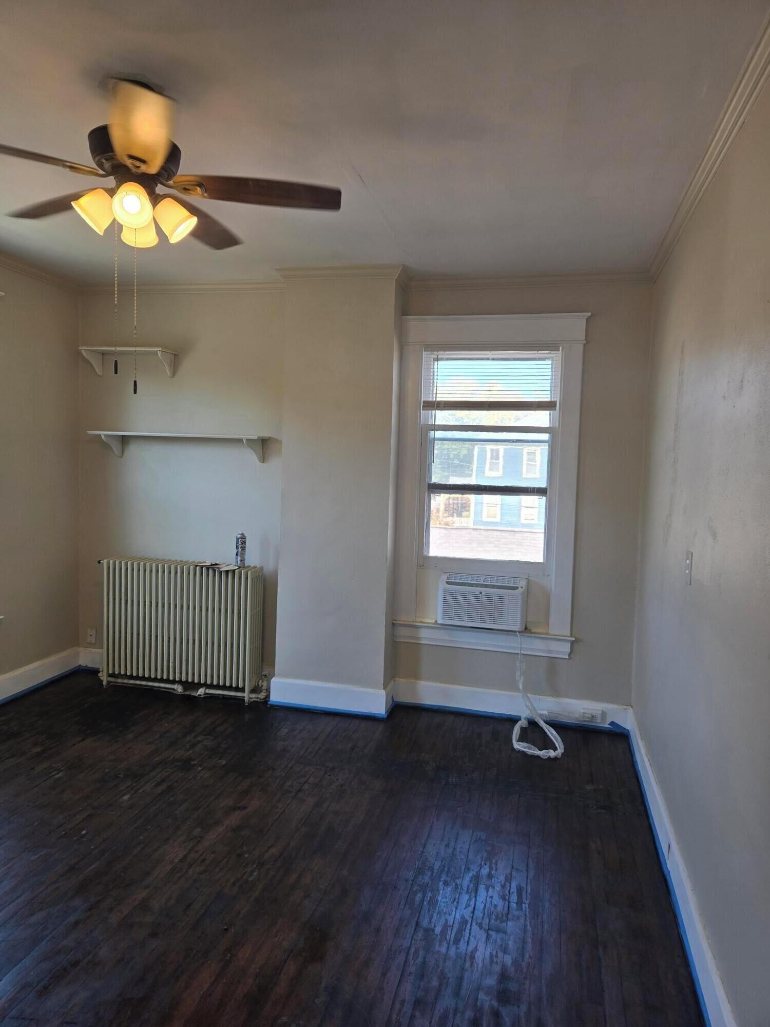 A room with a ceiling fan, radiator, window with blinds, and a radiator on the floor.