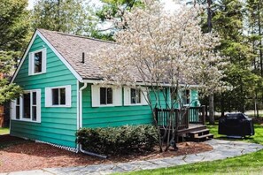 Una piccola casa a un piano, con esterno verde e decorazioni bianche, un portico con ringhiera in legno e un piccolo albero con fiori bianchi.