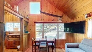 A wooden cabin interior with a dining table, chairs, a sofa, a TV, and a window overlooking a wooded area.