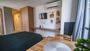 Et værelse med en seng, et træbord med en flettede bordskåner, et fjernsyn på væggen og en lille potteplante.