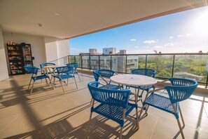 Ein Balkon mit blauen Stühlen und einem Tisch, ein Getränkeautomat und Blick auf Gebäude und Grünflächen.