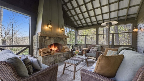 A cozy outdoor seating area with wicker furniture, a stone fireplace, and large windows offering a view of trees. - Sunset Nest, Walk to Town (Highlands)
