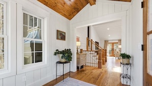 A spacious hallway with wooden flooring, white walls, and a wooden ceiling. There are two potted plants on stands, a staircase leading to another room, and a large window with multiple panes. - Sunset Nest, Walk to Town (Highlands)