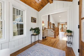 A spacious hallway with wooden flooring, white walls, and a wooden ceiling. There are two potted plants on stands, a staircase leading to another room, and a large window with multiple panes. - Aschmeyer's, Walk to Town (Highlands)