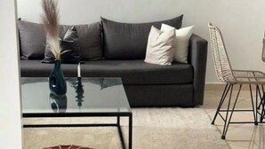 A modern living room with a dark sofa, a glass coffee table, a colorful pouf, and a wicker chair. - Nice apartment (Marrakech)