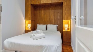 Uma cama bem arrumada com lençóis brancos e toalhas enroladas, painéis de madeira na parede e abajures laterais.