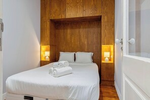 A neatly made bed with white linens and rolled towels, wooden paneling on the wall, and bedside lamps. - Live In Setúbal III (Setúbal)