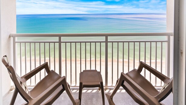 A balcony with two chairs and a small table, overlooking a beach with clear blue water and a distant shoreline. - Bundle of Three (3) Beachfront Penthouse Suites! Tons Of Resort Amenities! (Myrtle Beach)