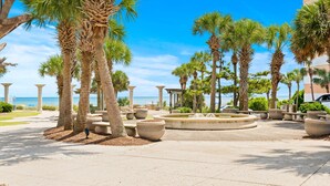Ein gepflasterter Weg, gesäumt von Palmen und großen Pflanzkübeln, führt zu einem Strand mit dem Meer im Hintergrund.