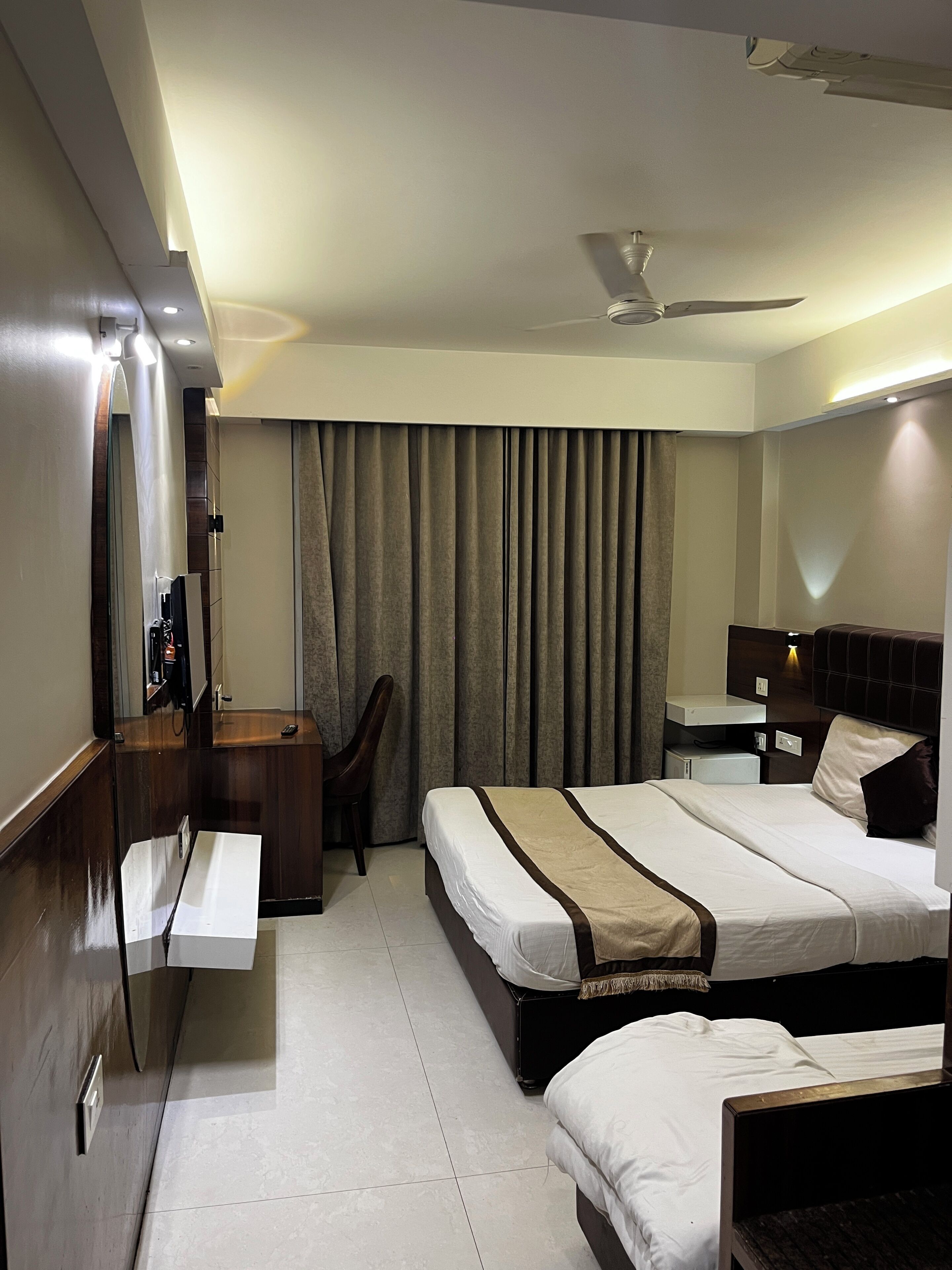 A hotel room with a bed, a desk, a chair, a mirror, a ceiling fan, and a window with curtains.